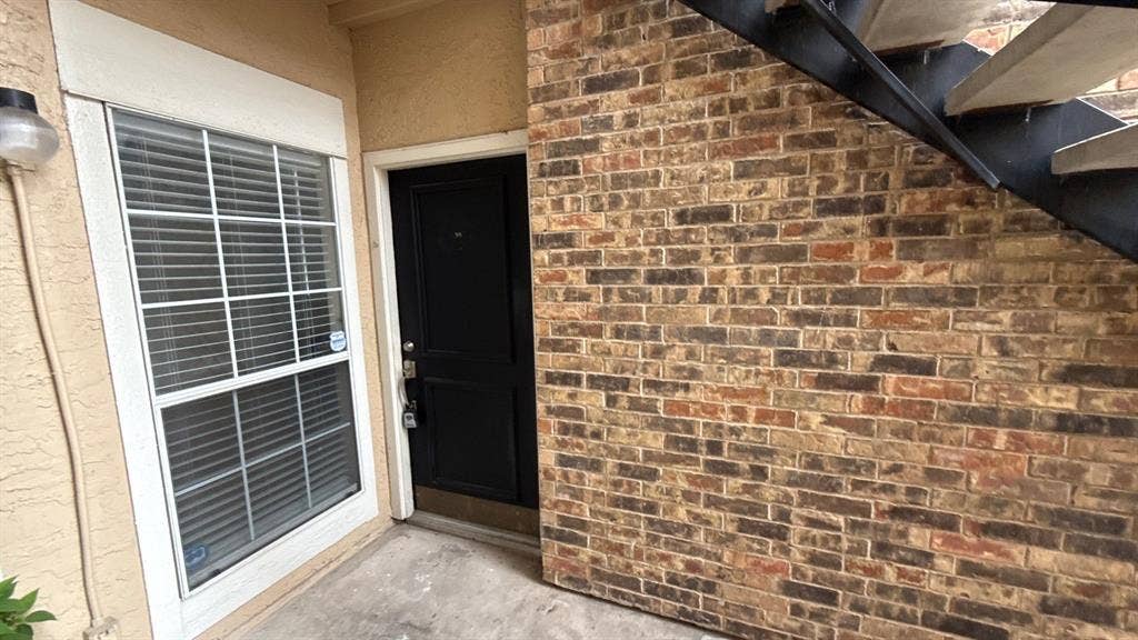 Exterior entryway featuring multi-tonal brick masonry, a solid black front door, a single-hung window with white trim, and a metal staircase