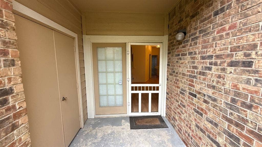Covered entryway featuring a brick facade, exterior storage closet, glass-panel entry door, and a screen door with a dome-style wall-mounted light fixture