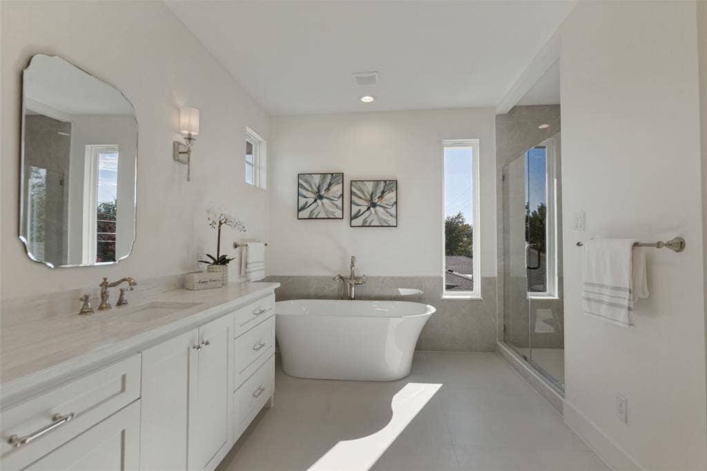 304 S Weatherred Drive, Richardson, TX 75080 Freestanding soaking tub with floor-mounted faucet, glass-enclosed shower, white vanity cabinet with stone-finish countertop, scalloped-edge wall mirror, and dual vertical windows