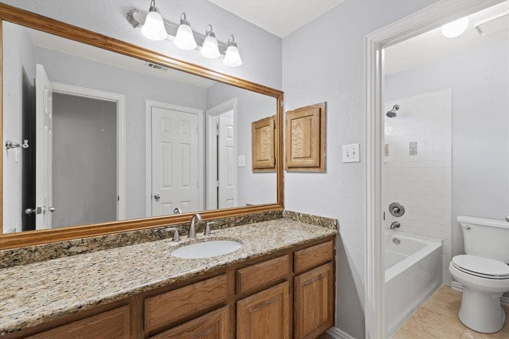 892 Brentwood Drive, Coppell, TX 75019 Upstairs 2nd Bathroom featuring a stone-look countertop, wood cabinetry, expansive wall-mounted mirror, four-light vanity fixture, and a tub-shower combination with white subway tile surround