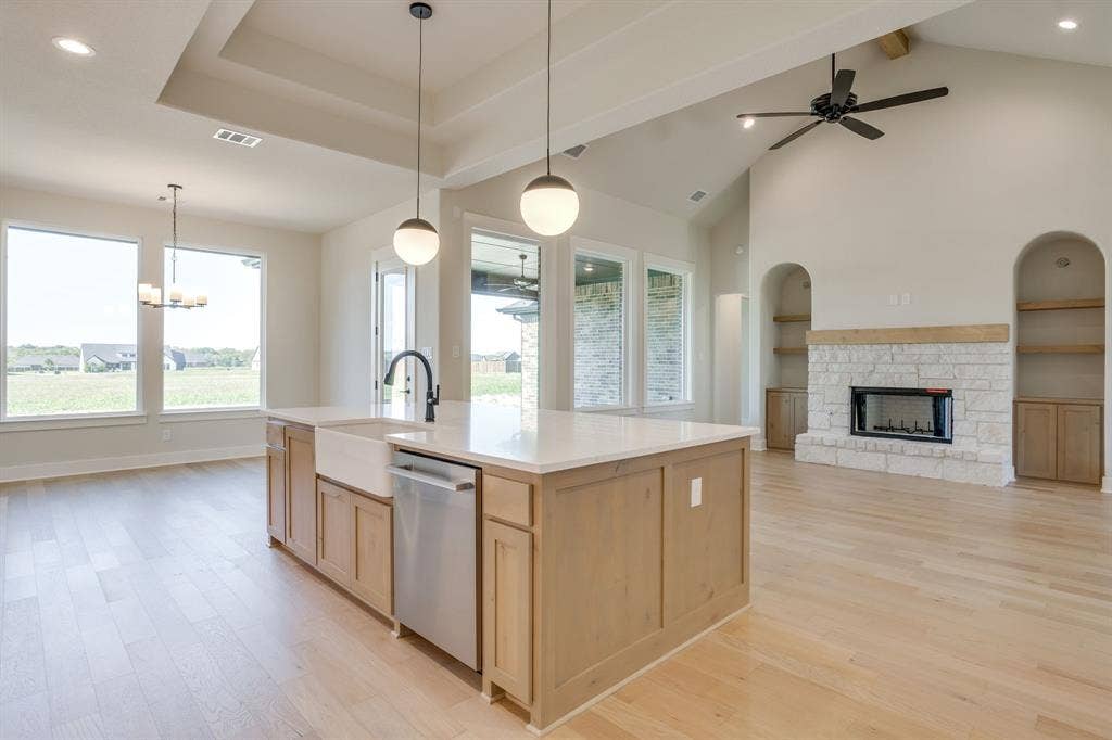 501 Olson Road, Waxahachie, TX 75167 Open-concept floor plan featuring light wood-finish flooring, a central kitchen island with a farmhouse sink and quartz countertops, vaulted ceilings, and a floor-to-ceiling stone fireplace flanked by built-in shelving