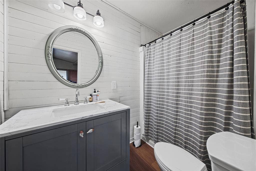 406 Summit Street, Farmersville, TX 75442 Dark gray vanity with a stone-top integrated sink, horizontal shiplap wall paneling, circular mirror with decorative frame, wood-finish flooring, and a three-light vanity fixture