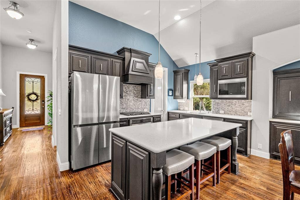 Kitchen featuring dark-stained cabinetry, a central island with a stone-composite countertop, stainless steel refrigerator, patterned tile backsplash, and vaulted ceilings