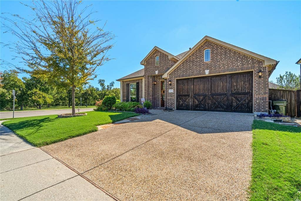 Brick exterior featuring an arched entryway, decorative dark wood-finish garage doors, exposed aggregate concrete driveway, and a manicured front lawn