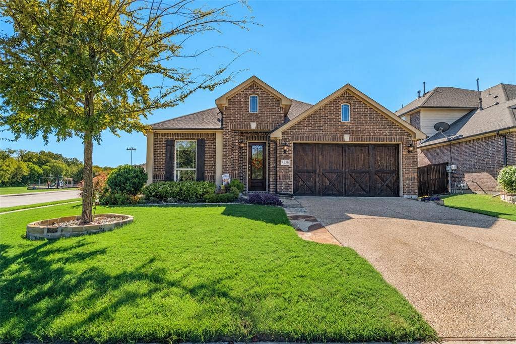 Brick exterior with a gabled roofline, decorative wood-finish garage door, and a manicured front lawn with a stone-bordered tree planter