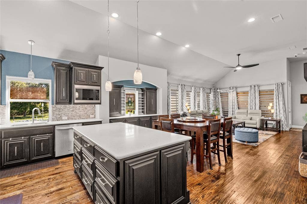 Open-concept space with vaulted ceilings, wood-finish flooring, dark-finish cabinetry, a large kitchen island with light-toned solid surface countertops, and recessed lighting