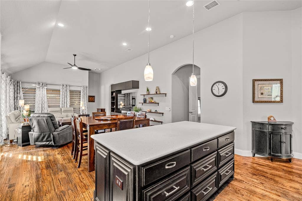 Vaulted ceiling featuring recessed lighting, a central kitchen island with a stone-finish countertop and dark cabinetry, and wood-finish flooring