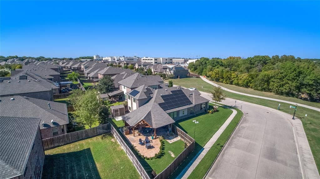 Aerial perspective of a multi-gabled roofline featuring integrated solar panels, a detached stone-paver patio with a covered structure, and a fenced yard with lawn