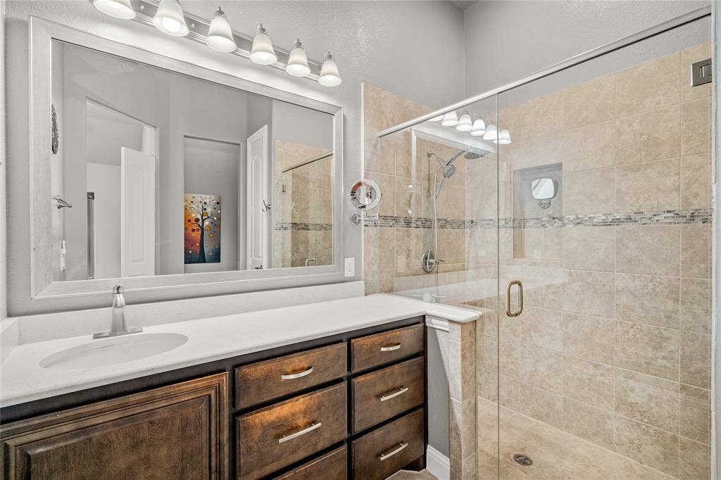 Bathroom featuring a wood-finish vanity with a solid-surface countertop, an expansive framed mirror, a multi-bulb vanity light, and a walk-in shower with tile surround and a glass door