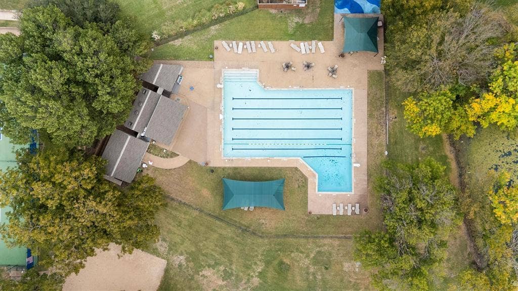 Aerial view of a swimming pool with multiple lap lanes, paved pool deck, shaded canopy structures, and mature tree-lined perimeter