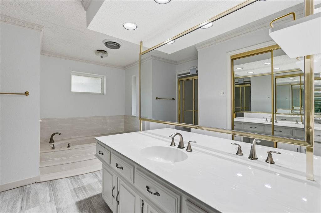 Dual-sink vanity with white stone countertop, integrated sinks, brass-trimmed wall mirror, built-in soaking tub with surround tile, and stone-finish flooring