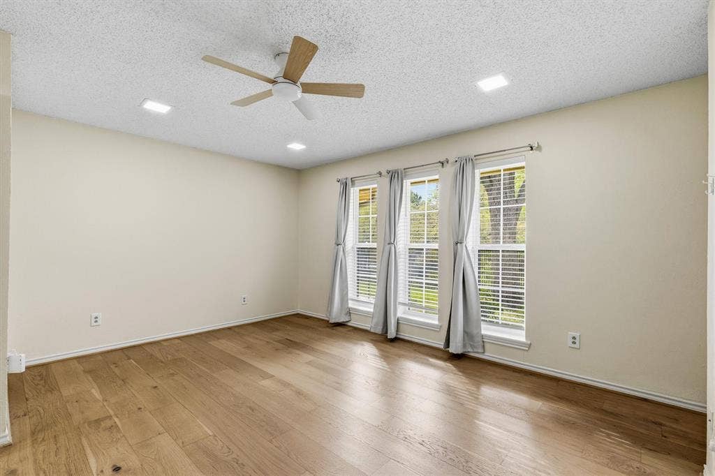 Room featuring wood-finish flooring, a multi-blade ceiling fan, and three windows with neutral-toned trim