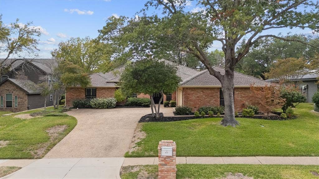 Single-story brick exterior featuring a multi-gabled roofline, an expansive concrete driveway, and established shade trees with landscaped garden beds and J swing garage