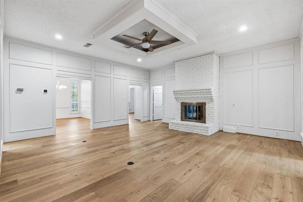 Living area featuring wood-finish flooring, white painted brick fireplace with hearth, recessed tray ceiling with ceiling fan, and extensive wall paneling