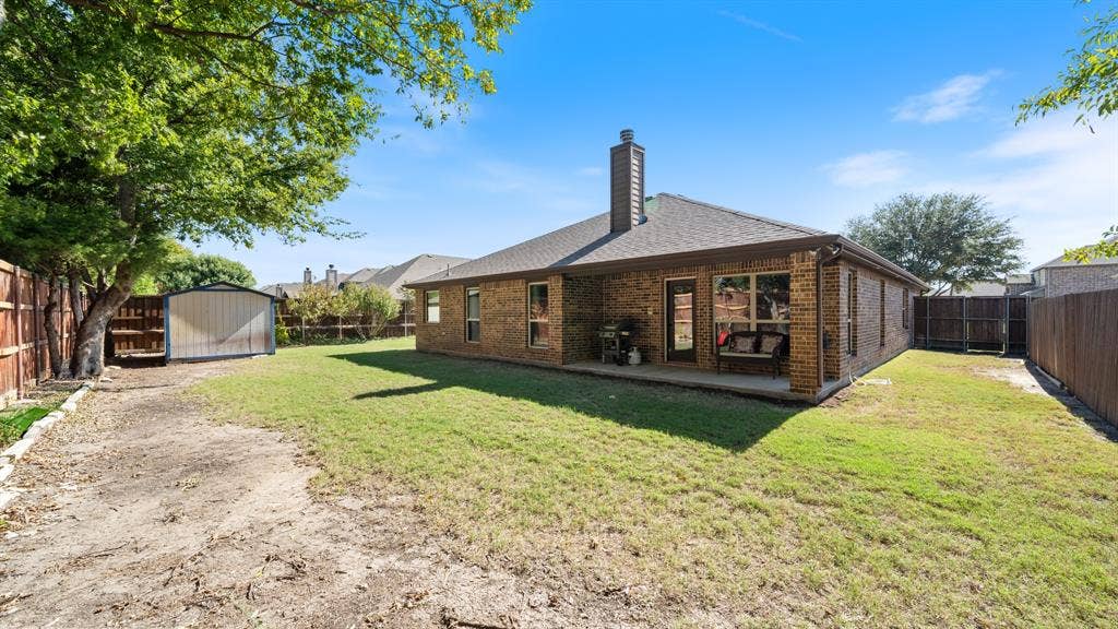 1802 Watermark Lane, Wylie, TX 75098 Backyard featuring a brick exterior, covered patio, masonry chimney, perimeter wood fencing, and a detached metal storage structure