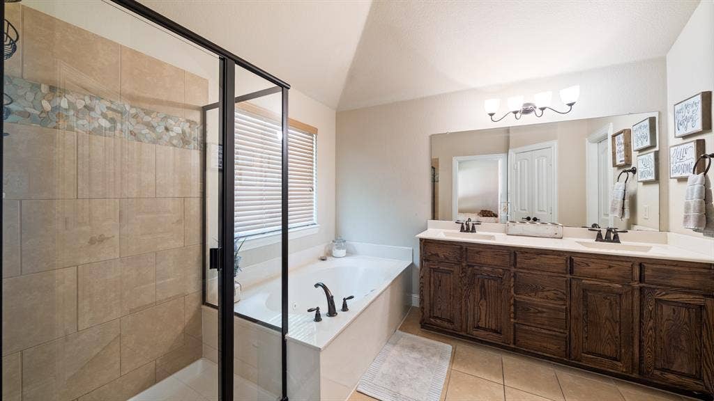 1802 Watermark Lane, Wylie, TX 75098 Dual vanity with dark wood-finish cabinetry, soaking tub, glass-enclosed shower with tiled walls and decorative stone inlay, and wall-mounted light fixture