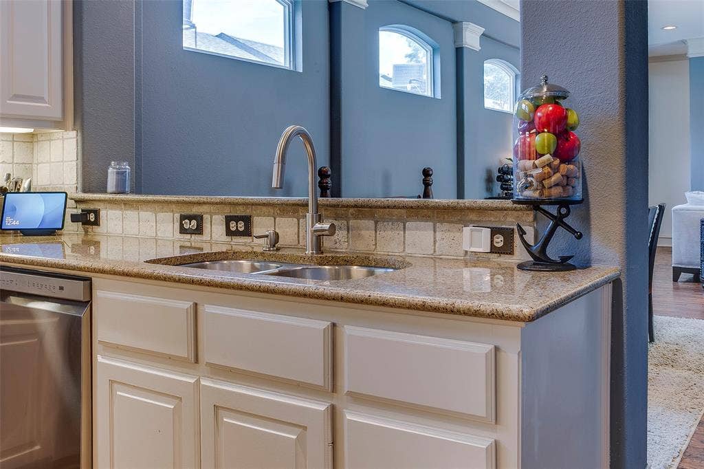 Kitchen featuring a double stainless steel sink with a gooseneck faucet, light granite countertops, white cabinetry with recessed panel doors, and a tiled backsplash