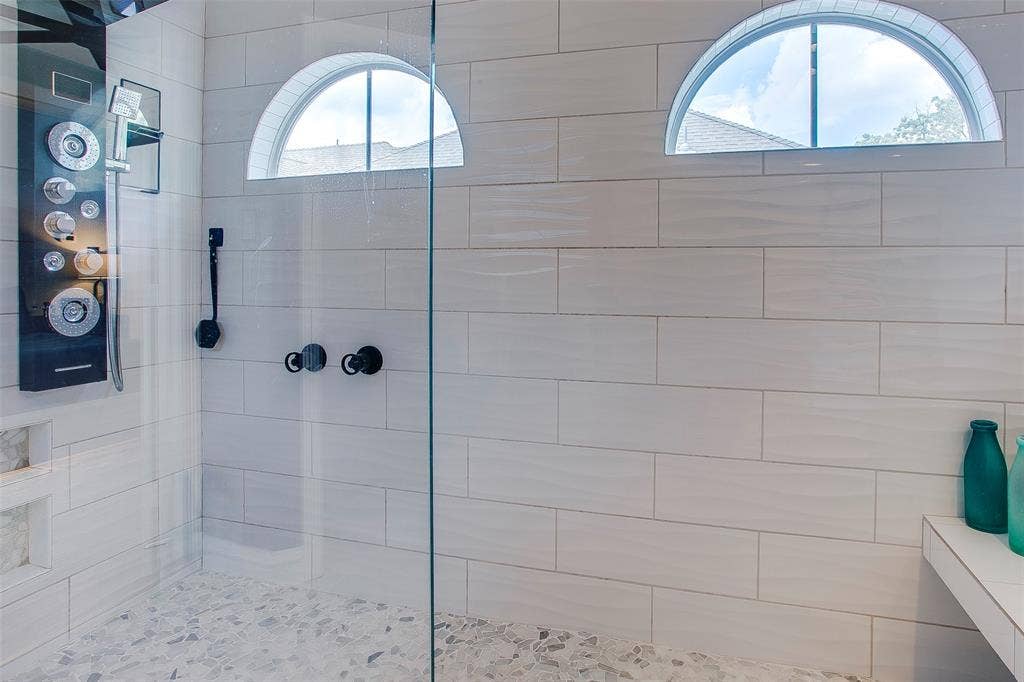 Glass-enclosed shower featuring a multi-function shower panel, two arched windows, a built-in bench, and pebble-tile flooring