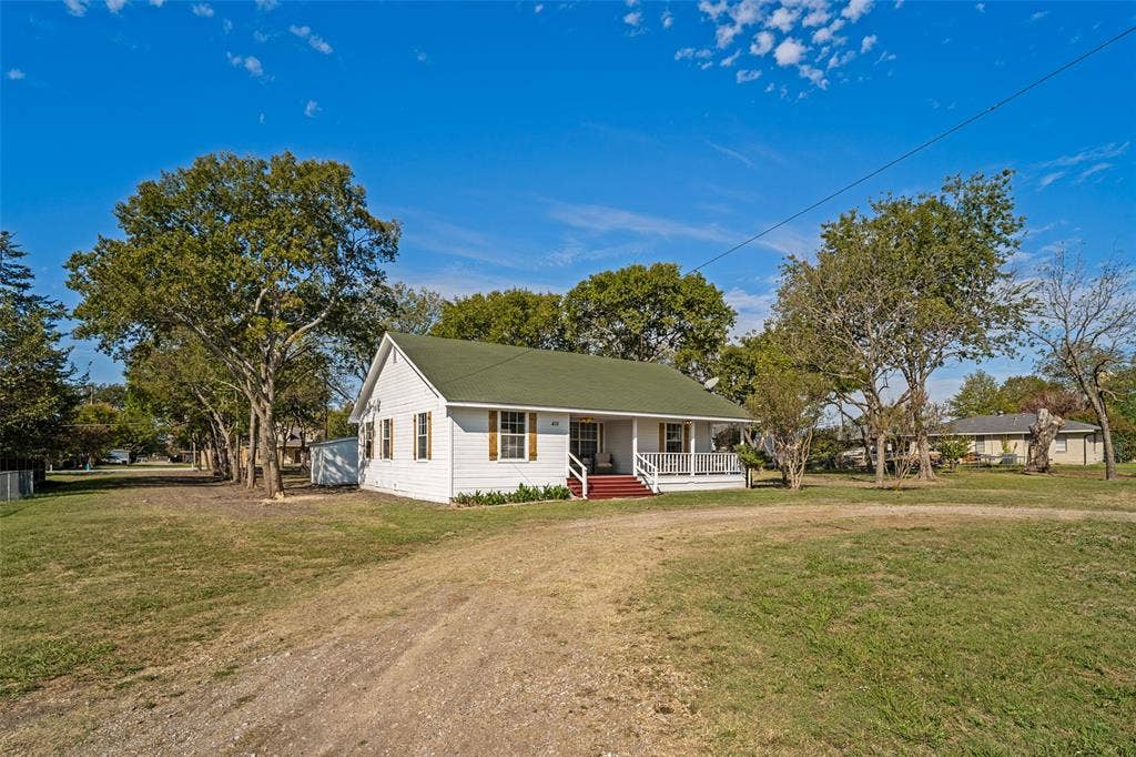 415 S Bell Street, Royse City, TX 75189 Exterior featuring white siding, a green gabled roof, an expansive lawn, mature trees, and a covered front porch with a wood-finish staircase