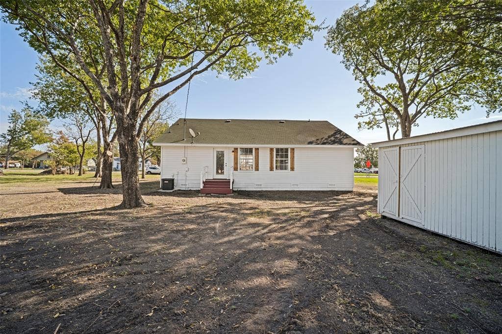 415 S Bell Street, Royse City, TX 75189 White exterior siding, shingle roofing, wooden window shutters, and a detached storage structure on a spacious lot with mature trees