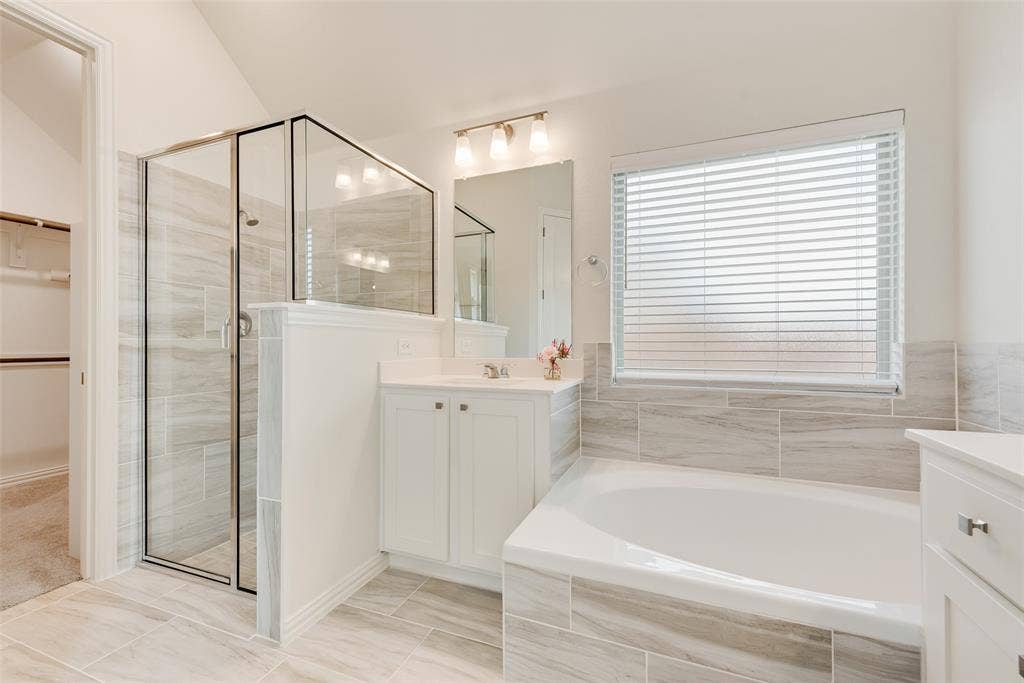 3934 Timberview Street, Midlothian, TX 76065 Bathroom featuring a walk-in glass-enclosed shower, soaking tub with tile surround, white cabinetry, rectangular mirror, and light-tone tile flooring