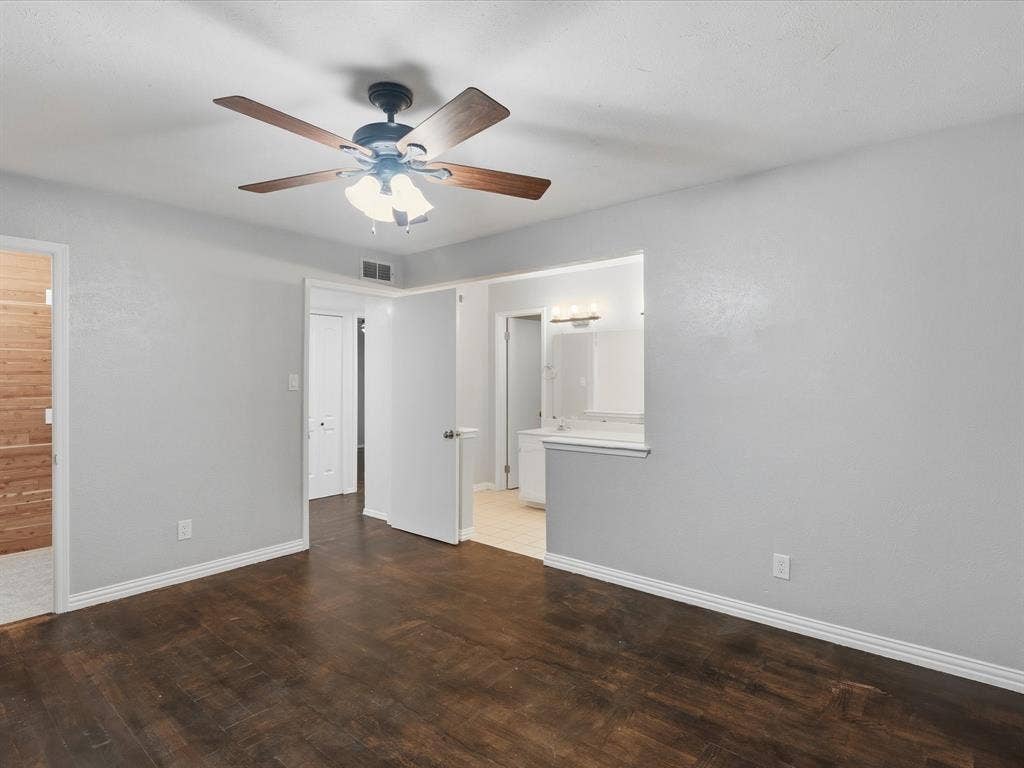 Room featuring wood-finish flooring, a ceiling fan with integrated lighting, neutral wall paint, and an open doorway revealing a vanity area