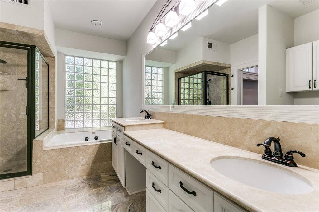 1214 Saint Regis Drive, Irving, TX 75038 Dual vanity featuring white cabinetry, stone-surface countertops, and dark hardware, complemented by a glass-block window, deep soaking tub, and a tiled walk-in shower