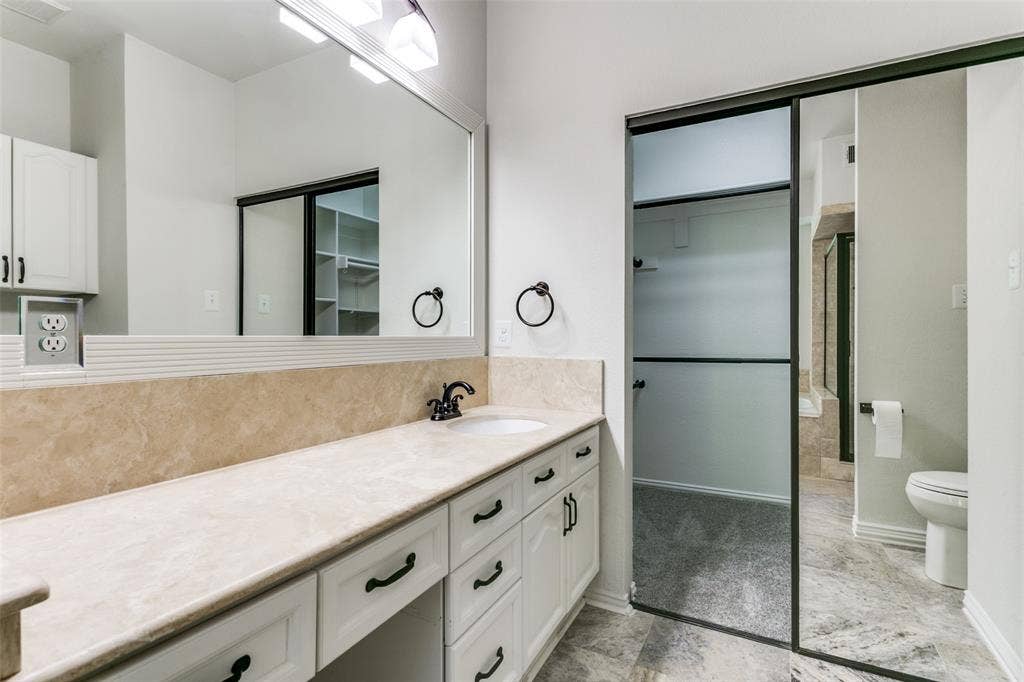 1214 Saint Regis Drive, Irving, TX 75038 Vanity featuring a stone countertop with an integrated basin, white cabinetry with dark hardware, a full-width mirror, and a sliding glass door