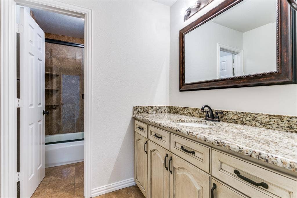 1214 Saint Regis Drive, Irving, TX 75038 Bathroom featuring stone-surface vanity with undermount sink, oil-rubbed bronze faucet, custom cabinetry with dark hardware, and a tile-surround shower with built-in shelving