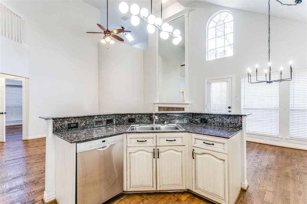 1214 Saint Regis Drive, Irving, TX 75038 Kitchen featuring an island with stone-finish countertops, stainless steel dishwasher, double basin undermount sink, and light-toned cabinetry, accented by dual overhead chandeliers and vaulted ceilings