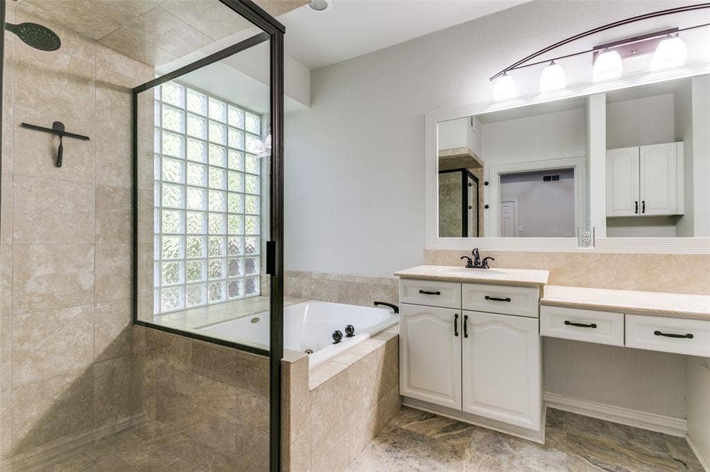 1214 Saint Regis Drive, Irving, TX 75038 Bathroom featuring a jetted soaking tub, glass-enclosed shower with tile surround, white cabinetry, neutral stone-tile flooring, and a glass block window