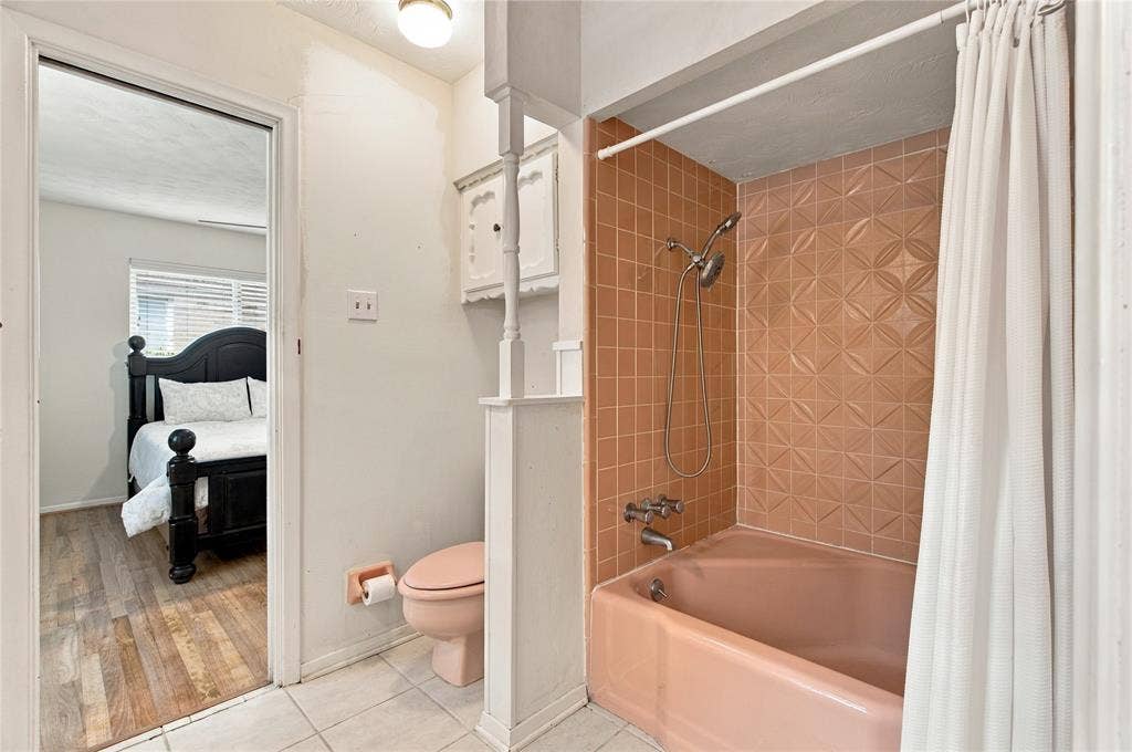 Bathroom featuring a tub with an integrated shower head and geometric-patterned tile surround, pink porcelain fixture, wall-mounted cabinetry, and tile flooring