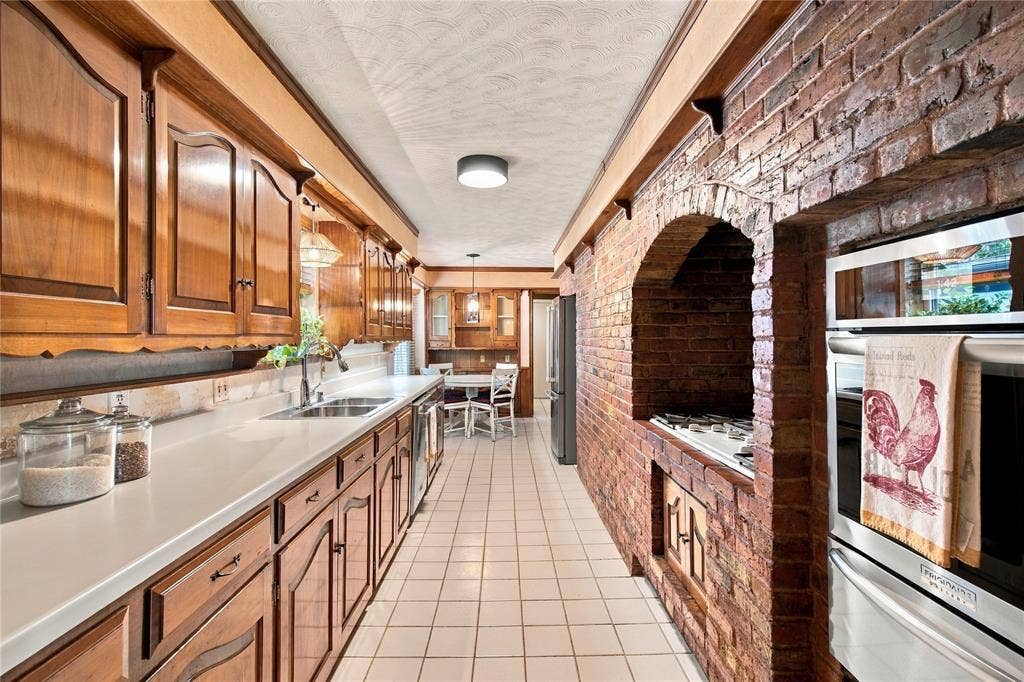 Galley kitchen featuring wood cabinetry, solid surface countertops, exposed brick wall with arched brick cooktop surround, and ceramic tile flooring