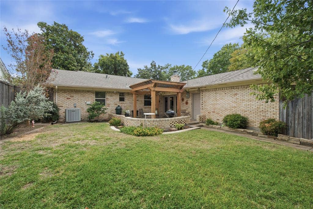 Brick exterior featuring a wooden pergola, raised masonry patio, manicured lawn, and perimeter wood fencing