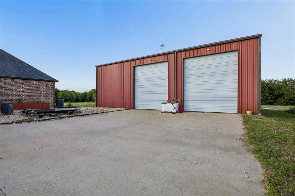 Detached metal workshop featuring dual roll-up garage doors, a concrete driveway, and exterior wall-mounted lighting