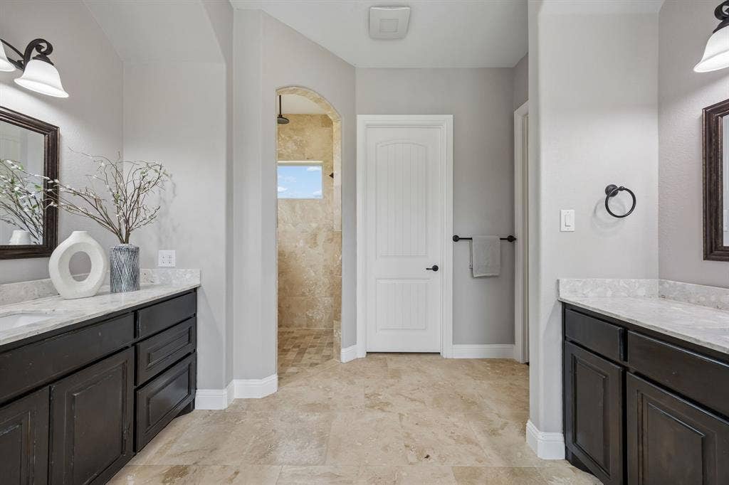 Dual dark-finish cabinetry with stone-surface countertops, stone tile flooring, arched shower entry, and wall-mounted vanity lighting