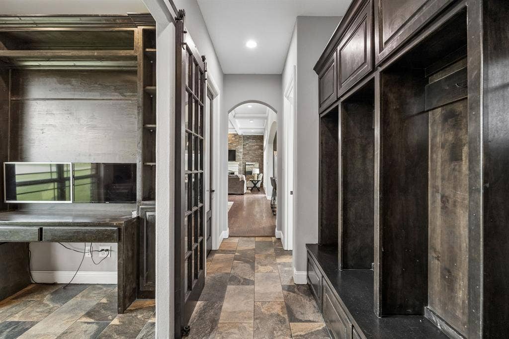 Built-in dark wood cabinetry with integrated desk space, custom mudroom lockers, sliding glass-paneled barn door, arched hallway entry, and stone-finish tile flooring