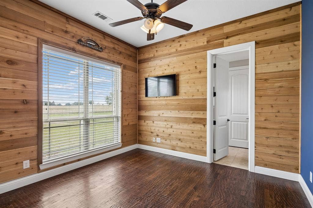 Room featuring horizontal wood-plank wall paneling, dark wood-finish flooring, a ceiling fan with integrated lighting, and a large window with horizontal blinds