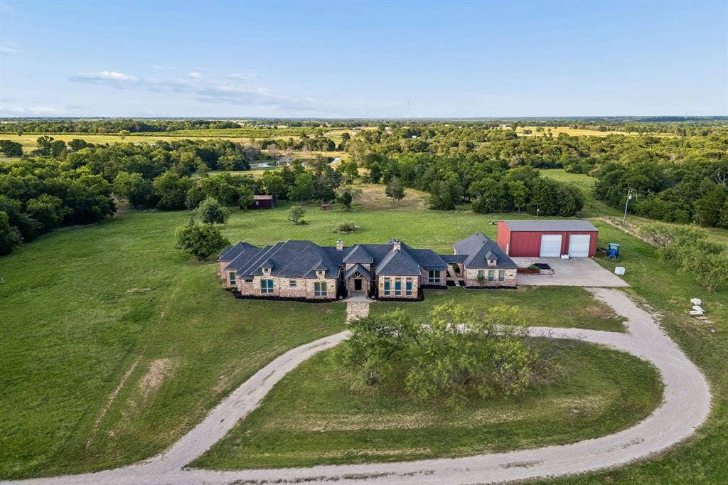 Aerial perspective of a multi-gabled stone and brick exterior, expansive acreage with mature trees, detached metal workshop with concrete driveway, and a gravel circular motor court