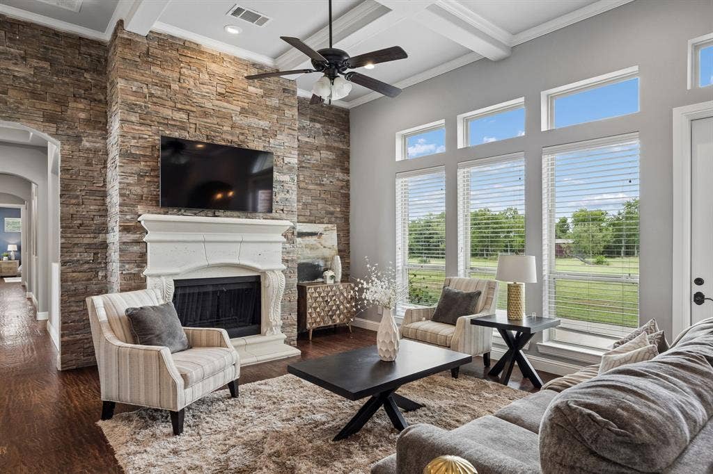 Living area featuring a floor-to-ceiling stacked stone fireplace, ornate mantle, coffered ceiling, tall windows with transom accents, and wood-finish flooring