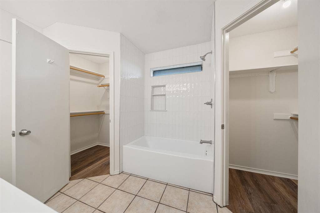 4043 Country Meadows Circle, Granbury, TX 76049 Bathroom featuring a white tile tub surround with built-in recessed shelving, clerestory window, metal shower fixture, ceramic tile flooring, and dual walk-in closets with wood-finish flooring