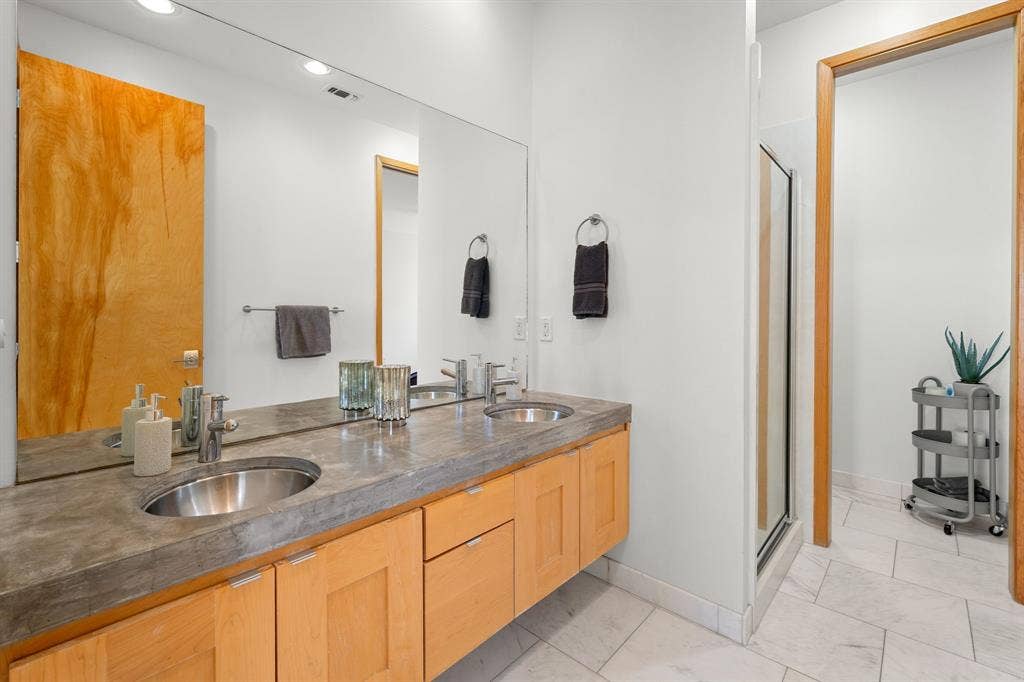 1997 Hollystone Drive, Tyler, TX 75703 Double vanity featuring natural wood cabinetry, integrated concrete-style countertop, dual stainless steel drop-in sinks, and a full-width wall mirror