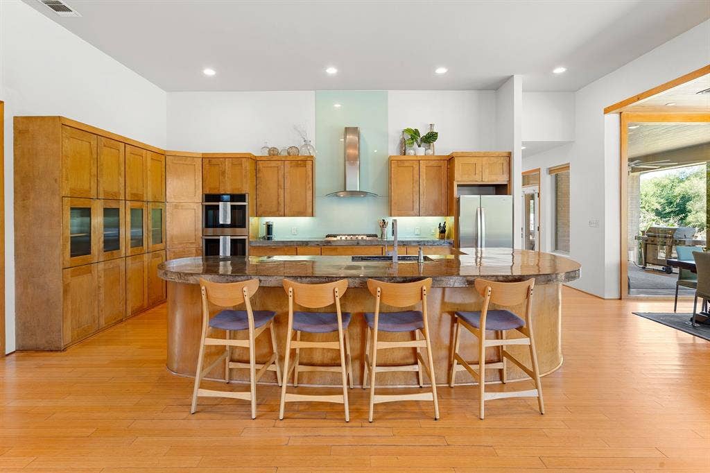 1997 Hollystone Drive, Tyler, TX 75703 Kitchen featuring wood cabinetry, curved stone-topped island with seating, stainless steel double wall ovens, stainless steel range hood, and wood-finish flooring