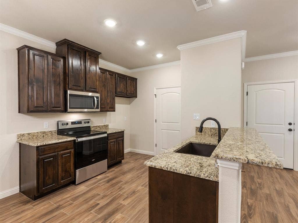 Kitchen featuring stained wood cabinetry, stone countertops, stainless steel appliances, a center island with a drop-in sink, and wood-finish flooring
