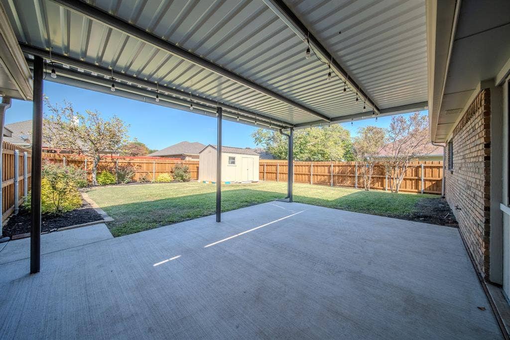 2213 Moore Drive, Plano, TX 75074 Covered concrete patio with a metal roof, wood-plank privacy fencing, and a detached storage structure set within a grass lawn