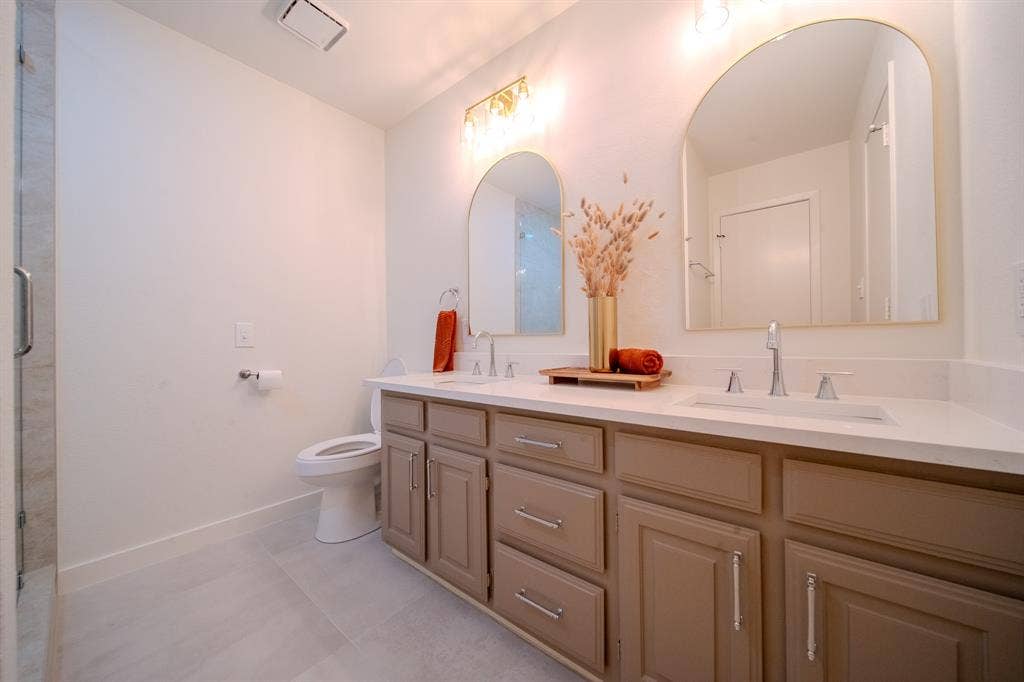 2213 Moore Drive, Plano, TX 75074 Dual-sink vanity with light-tone cabinetry, stone-surface countertop, arched wall-mounted mirrors, and chrome-finish fixtures