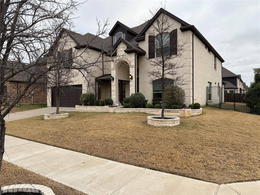 14518 Shady Grove Lane, Frisco, TX 75035 Two-story exterior featuring light-toned brick masonry, arched entryway, stacked stone retaining walls, and dark-finished decorative window shutters