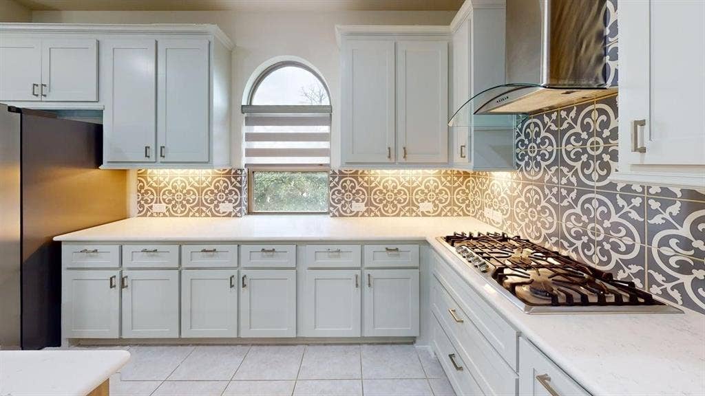 14518 Shady Grove Lane, Frisco, TX 75035 Kitchen featuring white shaker-style cabinetry, patterned tile backsplash, solid-surface countertops, gas cooktop with stainless steel range hood, and arched window