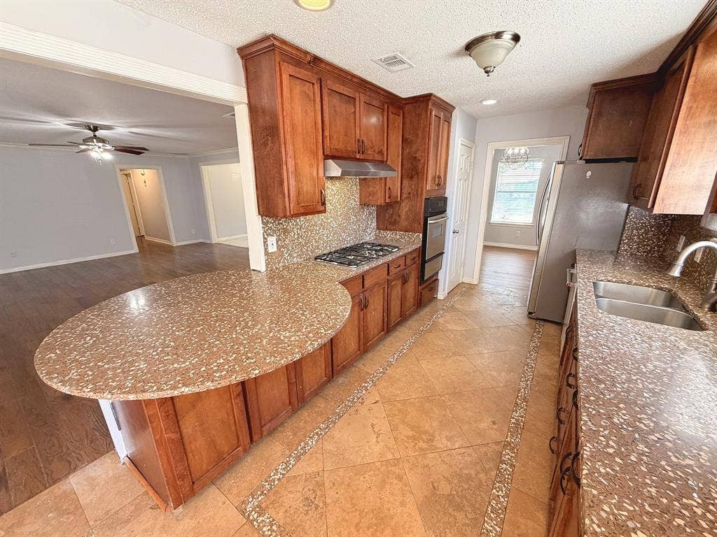 2701 Northcrest Drive, Plano, TX 75075 Kitchen featuring stained wood cabinetry, stone-composite countertops with a rounded breakfast bar, built-in stainless steel double wall oven, gas cooktop, and stone tile flooring