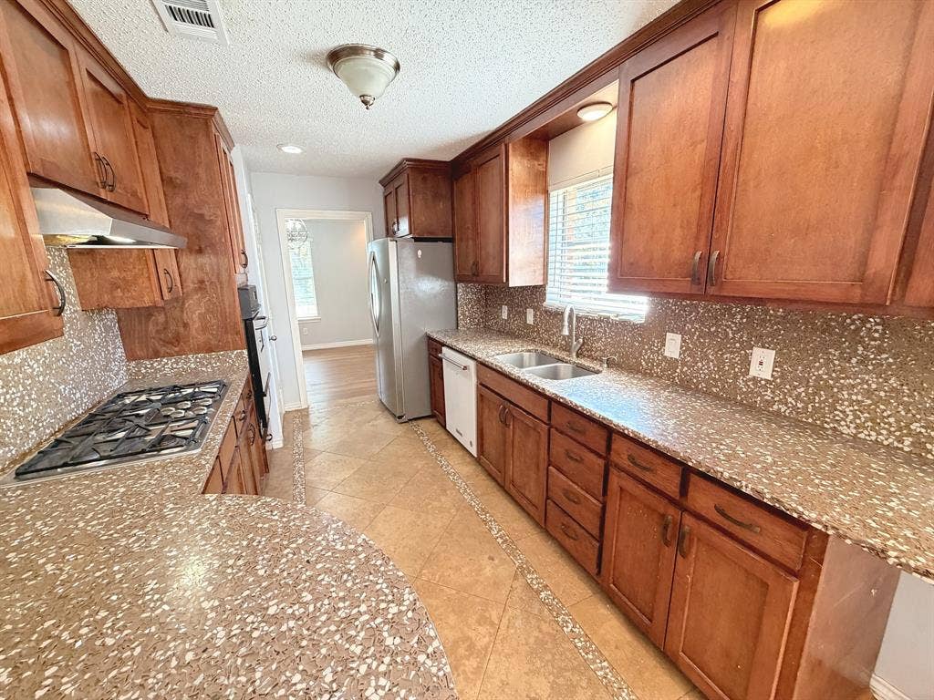 2701 Northcrest Drive, Plano, TX 75075 Kitchen featuring stained wood cabinetry, stone-style countertops, mosaic tile backsplash, integrated stainless steel gas cooktop, and tile flooring
